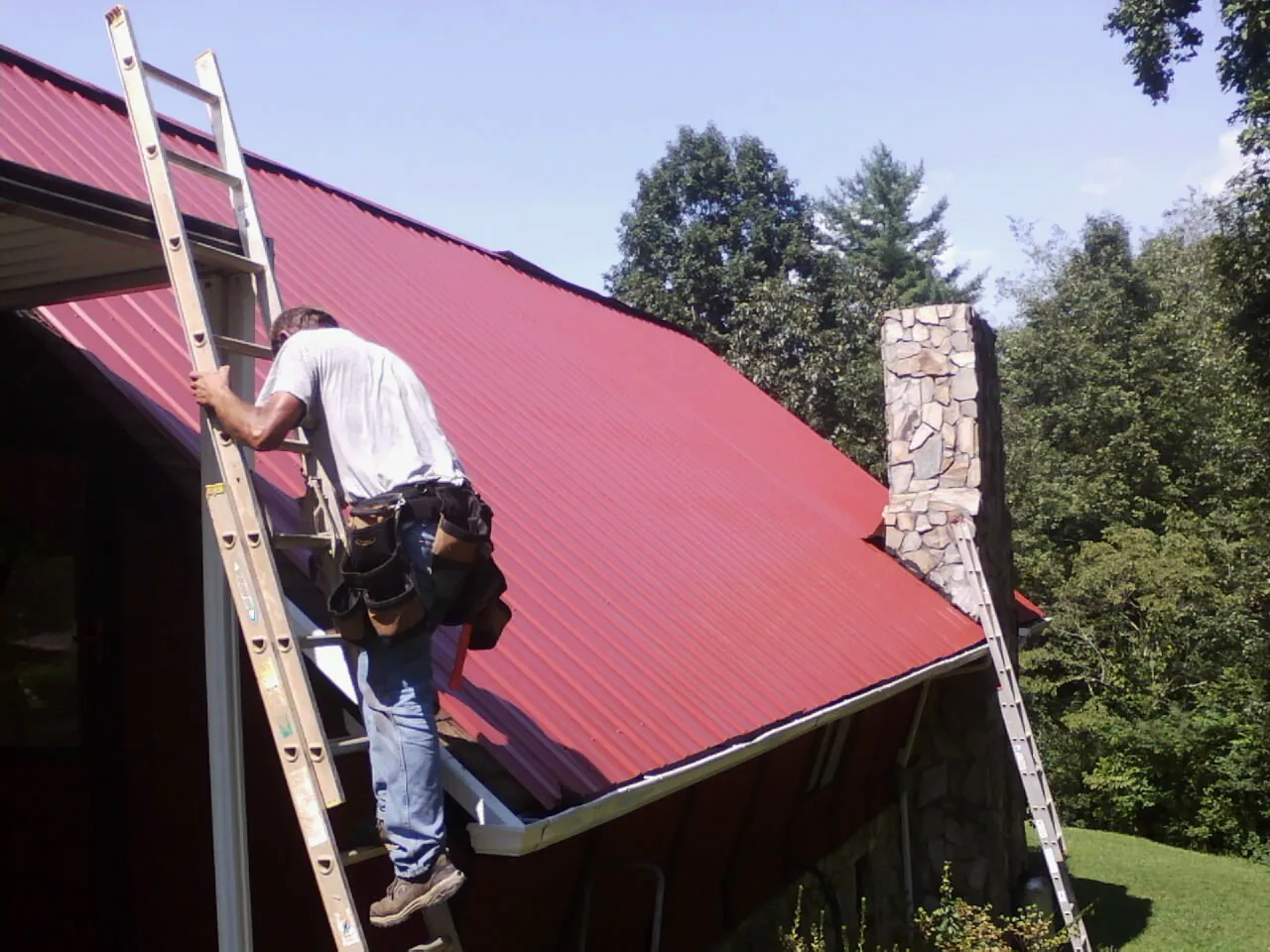 Roofer on a ladder beside a red metal roof and stone chimney — Soffit & Fascia Repair by Roofing Brothers in Vining